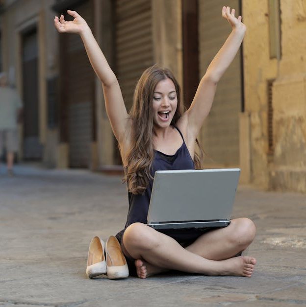 A cheerful woman sitting outdoors, celebrating success with arms raised, while using a laptop.
